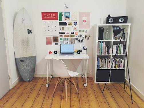 A neatly organized home office or study space featuring a small white desk with a laptop showing the time, a white chair, and various stationery items. On the wall, there is a calendar and an assortment of pictures and notes. A surfboard stands leaning against the wall on the left side. A white bookshelf on the right holds a collection of books, a pair of speakers on top, and a floor lamp illuminating the area.