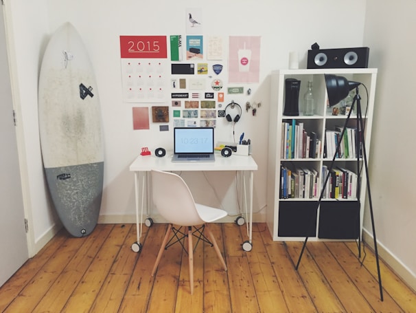 A neatly organized home office or study space featuring a small white desk with a laptop showing the time, a white chair, and various stationery items. On the wall, there is a calendar and an assortment of pictures and notes. A surfboard stands leaning against the wall on the left side. A white bookshelf on the right holds a collection of books, a pair of speakers on top, and a floor lamp illuminating the area.