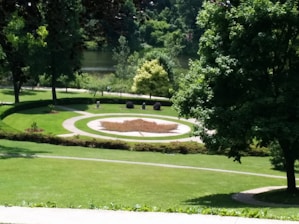 A landscaped garden area with a circular pathway surrounding a design featuring a maple leaf made from red-brown foliage. The garden is lush with green grass and surrounded by various trees and shrubs. In the background, a small body of water is visible, enhancing the serene natural setting.