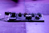 A stage or studio setup with an array of guitar effect pedals on a pedalboard. The pedals are connected with cables, and the lighting casts a bluish-purple hue over the scene.