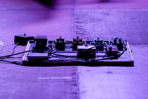 A stage or studio setup with an array of guitar effect pedals on a pedalboard. The pedals are connected with cables, and the lighting casts a bluish-purple hue over the scene.