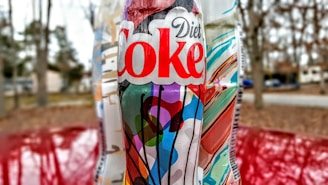 A Diet Coke bottle with a colorful, abstract label featuring various shapes and colors is in focus. The background is slightly blurred, showing an outdoor scene with trees and a roadway.