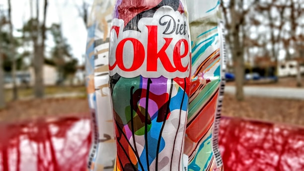 A Diet Coke bottle with a colorful, abstract label featuring various shapes and colors is in focus. The background is slightly blurred, showing an outdoor scene with trees and a roadway.
