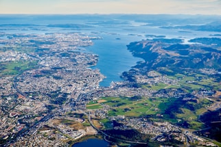 An aerial view of a coastal city surrounded by a large body of water and numerous islands. The landscape includes densely populated urban areas, sprawling green fields, and mountainous regions. The ocean inlets and bays create a complex coastline.