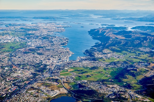 An aerial view of a coastal city surrounded by a large body of water and numerous islands. The landscape includes densely populated urban areas, sprawling green fields, and mountainous regions. The ocean inlets and bays create a complex coastline.
