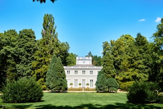 A grand, white classical-style mansion is set amidst a lush, green landscaped garden. Tall trees surround the property, and neatly trimmed shrubs and a manicured lawn enhance the elegant setting. The building features large windows and a symmetrical design, capped with a simple, elegant roof.