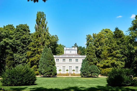 A grand, white classical-style mansion is set amidst a lush, green landscaped garden. Tall trees surround the property, and neatly trimmed shrubs and a manicured lawn enhance the elegant setting. The building features large windows and a symmetrical design, capped with a simple, elegant roof.