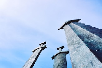 Three large stone swords are embedded into the ground, towering upwards against a clear blue sky. The perspective from below emphasizes their monumental and dramatic appearance.