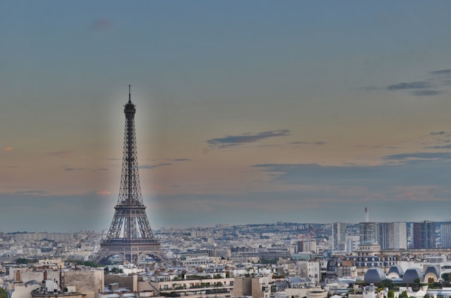 A panoramic view of a cityscape featuring the Eiffel Tower prominently against a twilight sky. Buildings extend across the foreground, with a mixture of modern and classic architecture.