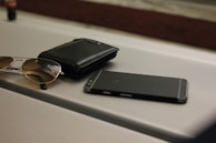 A pair of sunglasses, a black wallet, and a smartphone are placed on a ribbed surface, possibly a table or suitcase. The arrangement is casual, suggesting everyday personal items.