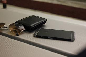 A pair of sunglasses, a black wallet, and a smartphone are placed on a ribbed surface, possibly a table or suitcase. The arrangement is casual, suggesting everyday personal items.