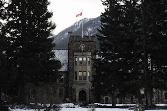 A stone building with a tower topped by a Canadian flag stands among large evergreen trees with a snow-covered mountain in the background.