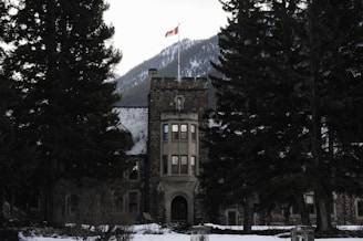 A stone building with a tower topped by a Canadian flag stands among large evergreen trees with a snow-covered mountain in the background.