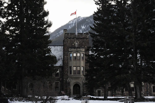 A stone building with a tower topped by a Canadian flag stands among large evergreen trees with a snow-covered mountain in the background.
