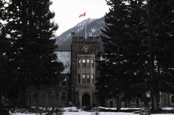 A stone building with a tower topped by a Canadian flag stands among large evergreen trees with a snow-covered mountain in the background.