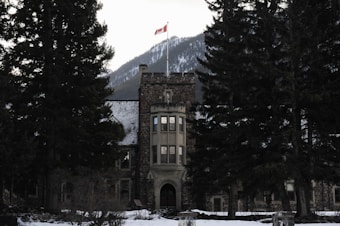 A stone building with a tower topped by a Canadian flag stands among large evergreen trees with a snow-covered mountain in the background.