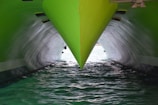 A close-up view of a green ship's bow cutting through the water, with two hulls creating a reflective effect on the water. The lighting highlights the sleek and polished surface of the vessel, contrasting with the darker, shadowed area below.