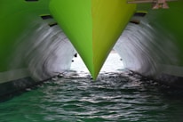 A close-up view of a green ship's bow cutting through the water, with two hulls creating a reflective effect on the water. The lighting highlights the sleek and polished surface of the vessel, contrasting with the darker, shadowed area below.