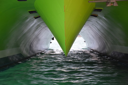 A close-up view of a green ship's bow cutting through the water, with two hulls creating a reflective effect on the water. The lighting highlights the sleek and polished surface of the vessel, contrasting with the darker, shadowed area below.