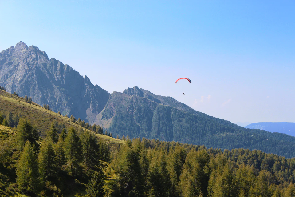 The image depicts a serene mountain landscape with a clear blue sky and lush green trees below. A paraglider is seen soaring high above, attached to a parachute that is visible in the top right corner of the frame. The mountains in the background are covered in dense forest, adding depth and texture to the scene.
