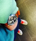 A close-up of a wristwatch with a brown leather strap and a detailed black dial is visible. The person wearing the watch is dressed in a green ribbed sweater and blue jeans. The floor tiles are dark, and the shoes have white bodies with distinct red accents on the laces.
