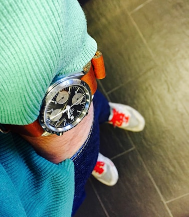 A close-up of a wristwatch with a brown leather strap and a detailed black dial is visible. The person wearing the watch is dressed in a green ribbed sweater and blue jeans. The floor tiles are dark, and the shoes have white bodies with distinct red accents on the laces.