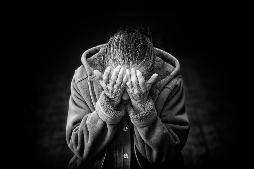 An old woman wearing a hoody coat and covering her face with her hands in yuri ria in black and white.