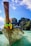 A traditional wooden boat with colorful decorations on the bow is anchored in crystal clear turquoise waters. Behind it are lush, green limestone cliffs under a bright blue sky with scattered clouds. The boat features writing on its side and a Thai flag is visible, indicating a tropical setting.