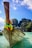 A traditional wooden boat with colorful decorations on the bow is anchored in crystal clear turquoise waters. Behind it are lush, green limestone cliffs under a bright blue sky with scattered clouds. The boat features writing on its side and a Thai flag is visible, indicating a tropical setting.