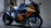 A sleek, modern motorcycle with a glossy orange and black finish is parked on a gravel surface near a wall. The design features sharp angles and aerodynamic elements, with the brand name visible on the side. The motorcycle appears well-maintained, with shiny components and intricate detailing on the body.