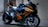 A sleek, modern motorcycle with a glossy orange and black finish is parked on a gravel surface near a wall. The design features sharp angles and aerodynamic elements, with the brand name visible on the side. The motorcycle appears well-maintained, with shiny components and intricate detailing on the body.