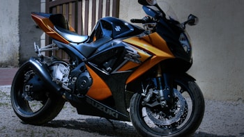 A sleek, modern motorcycle with a glossy orange and black finish is parked on a gravel surface near a wall. The design features sharp angles and aerodynamic elements, with the brand name visible on the side. The motorcycle appears well-maintained, with shiny components and intricate detailing on the body.