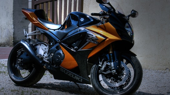 A sleek, modern motorcycle with a glossy orange and black finish is parked on a gravel surface near a wall. The design features sharp angles and aerodynamic elements, with the brand name visible on the side. The motorcycle appears well-maintained, with shiny components and intricate detailing on the body.