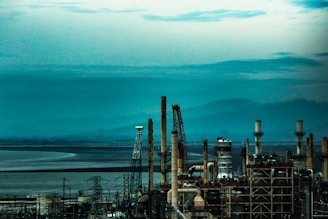 An industrial facility with numerous tall chimneys and towers set against a backdrop of mountains and a large body of water under a cloudy sky.