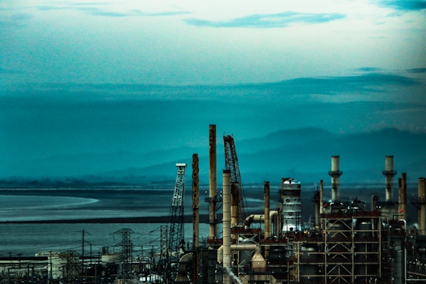 An industrial facility with numerous tall chimneys and towers set against a backdrop of mountains and a large body of water under a cloudy sky.