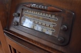 A vintage radio with a classic design featuring a large tuning dial and various frequency numbers. The radio has a wooden casing and multiple control knobs. It displays a variety of international radio stations, indicative of its age and style.