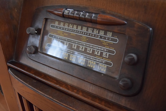 A vintage radio with a classic design featuring a large tuning dial and various frequency numbers. The radio has a wooden casing and multiple control knobs. It displays a variety of international radio stations, indicative of its age and style.