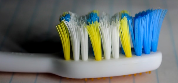 A close-up view of a toothbrush head with bristles in yellow, white, and blue colors. The toothbrush handle is white, and the background is blurred, giving emphasis to the bristles.