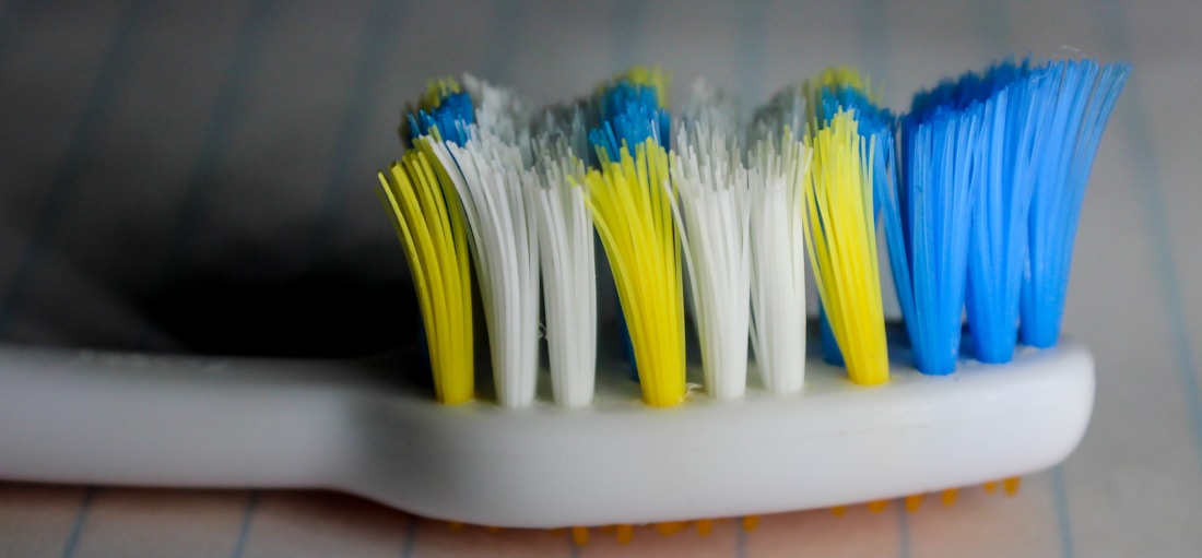 A close-up view of a toothbrush head with bristles in yellow, white, and blue colors. The toothbrush handle is white, and the background is blurred, giving emphasis to the bristles.