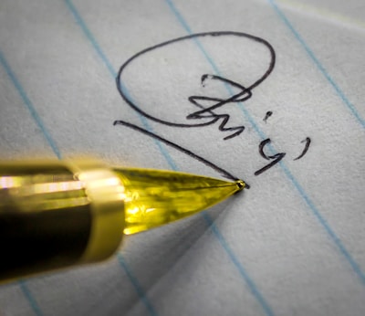 A close-up view of a pen tip positioned on a piece of lined paper with a signature partially visible. The pen's golden color contrasts with the paper's blue lines.