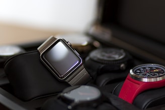 A collection of watches displayed in a watch case. The focus is on a modern digital watch with a metallic band and blank screen, positioned prominently. Surrounding it are various analog watches with different designs, including one with a red strap and detailed chronograph face.
