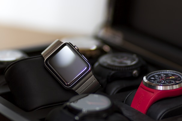 A collection of watches displayed in a watch case. The focus is on a modern digital watch with a metallic band and blank screen, positioned prominently. Surrounding it are various analog watches with different designs, including one with a red strap and detailed chronograph face.