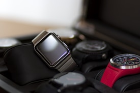 A collection of watches displayed in a watch case. The focus is on a modern digital watch with a metallic band and blank screen, positioned prominently. Surrounding it are various analog watches with different designs, including one with a red strap and detailed chronograph face.