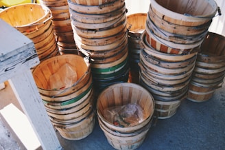 A collection of wooden baskets stacked neatly in piles. The baskets are made of light-colored wood with visible grain, and some have green bands around them. The setting appears to be outdoors with sunlight casting shadows, and there is a white wooden structure partially visible on the left.