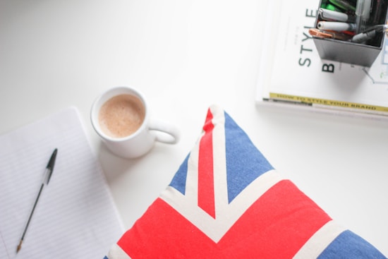 A workspace setup featuring a notebook with a pen, a coffee cup filled with a warm beverage, a book titled 'Style', and a Union Jack patterned pillow. The scene is organized on a white surface, creating a clean and aesthetically pleasing composition.