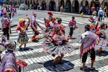 A group of people engaged in a traditional dance in a public square, wearing vibrant and colorful traditional attire featuring intricate patterns and designs. The dancers are energetically moving, and the background shows a cityscape with onlookers and colonial architecture.