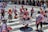 A group of people engaged in a traditional dance in a public square, wearing vibrant and colorful traditional attire featuring intricate patterns and designs. The dancers are energetically moving, and the background shows a cityscape with onlookers and colonial architecture.