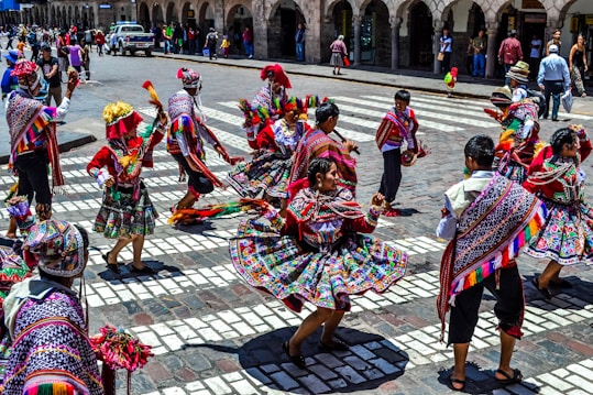 A group of people engaged in a traditional dance in a public square, wearing vibrant and colorful traditional attire featuring intricate patterns and designs. The dancers are energetically moving, and the background shows a cityscape with onlookers and colonial architecture.