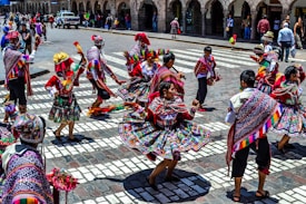A group of people engaged in a traditional dance in a public square, wearing vibrant and colorful traditional attire featuring intricate patterns and designs. The dancers are energetically moving, and the background shows a cityscape with onlookers and colonial architecture.