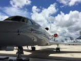 A large, sleek private jet is parked on a tarmac under a bright blue sky with scattered white clouds. Other smaller aircraft are visible in the background, and an airport control tower can be seen in the distance.